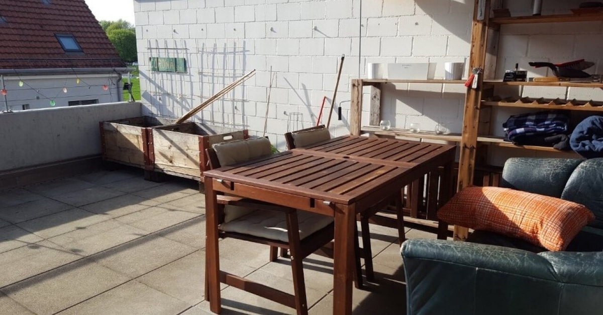 An outdoor balcony area features a wooden dining table surrounded by cushioned chairs. A green sofa is positioned nearby, providing a comfortable seating option. A planter box is visible along with gardening tools leaning against the wall. The space is illuminated by natural light.