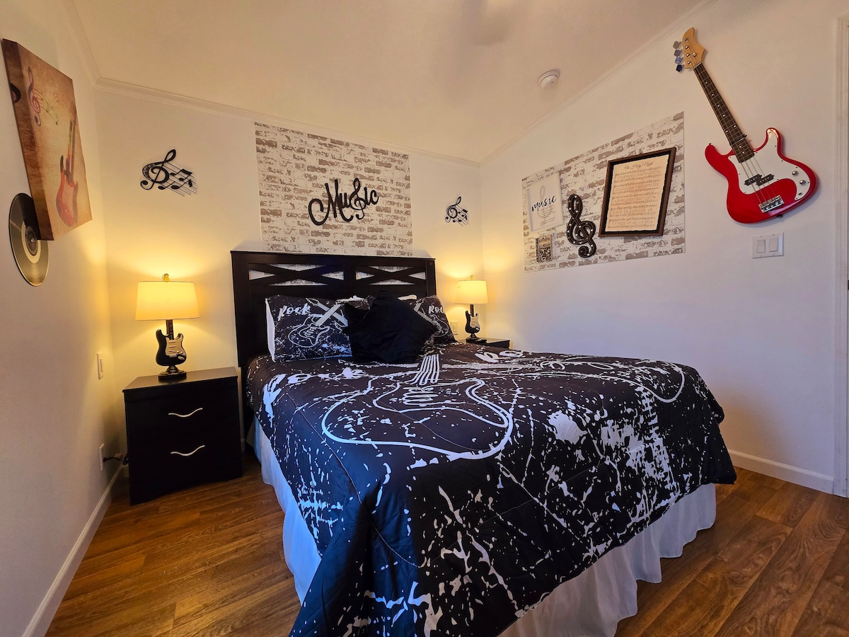 A cozy bedroom features a queen bed dressed in a black and white patterned comforter. Soft lighting is provided by two bedside lamps on dark wooden nightstands. Music-themed decor adorns the walls, while wooden flooring adds warmth to the space.