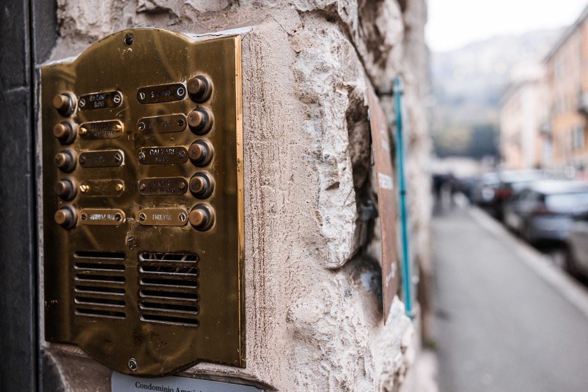 A brass intercom panel is mounted on a textured stone wall, featuring multiple buttons for different apartments. The panel reflects light subtly, while the cobblestone street and parked cars are visible in the background, suggesting a vibrant neighborhood.