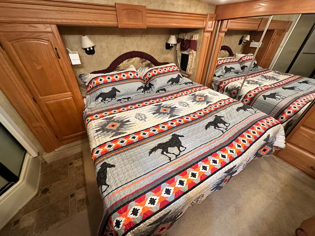 A cozy sleeping area features a quilted bedspread with a horse motif, complemented by warm wood cabinetry. Mirrored closet doors reflect the inviting decor, while soft lighting is provided by wall-mounted sconces on either side of the bed.
