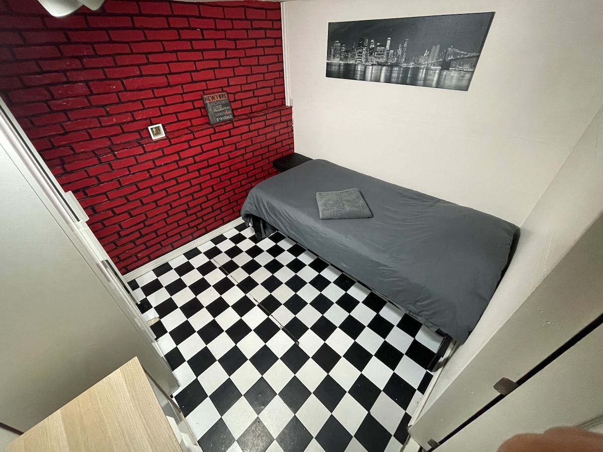 A minimalist bedroom displays a twin-size bed covered with a gray blanket, positioned against a red brick accent wall. The floor features a black and white checkered pattern, contributing to the room's unique character. A city skyline artwork is mounted above the bed.