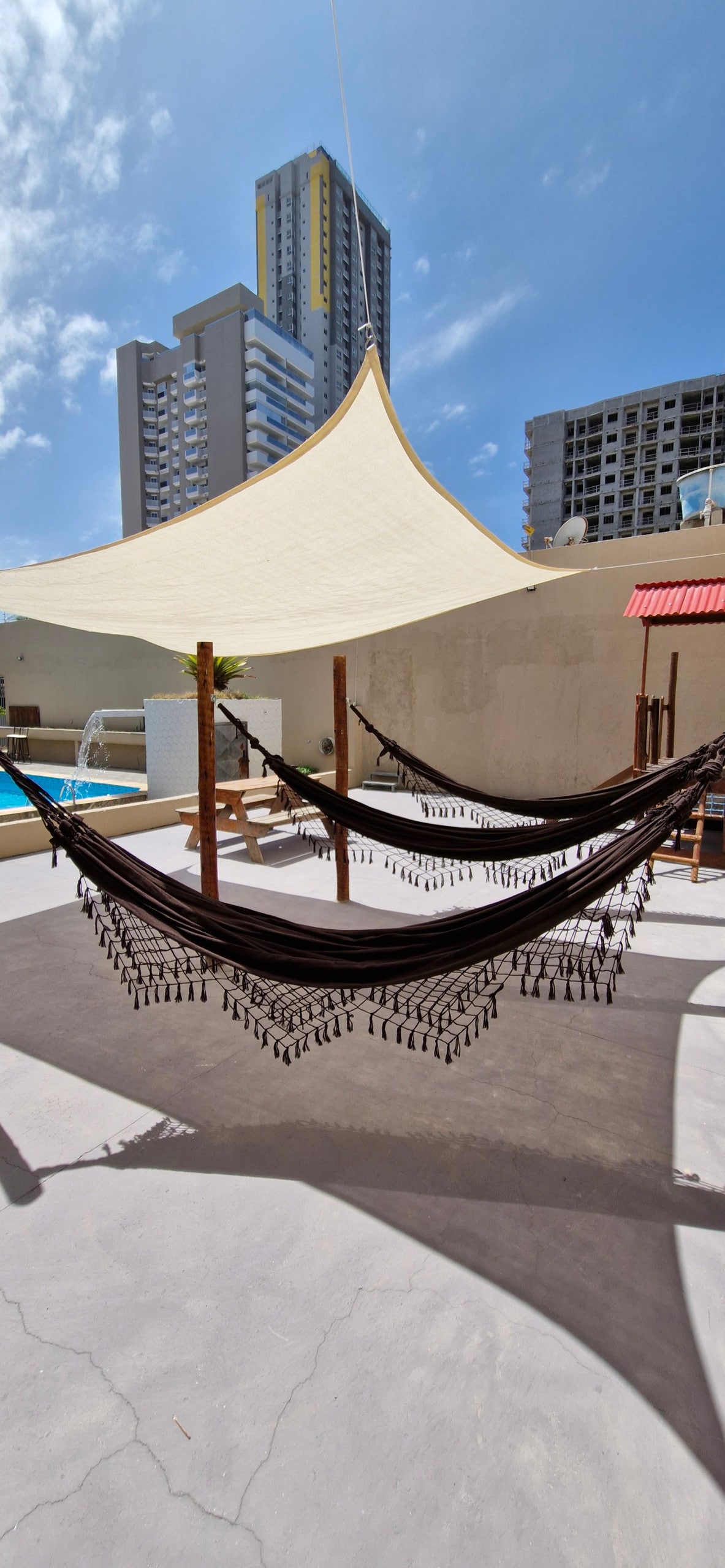 Two hammocks are suspended between wooden posts, providing a relaxing outdoor space. A large shade sail above offers protection from the sun. The pool area is visible in the background, with modern buildings framing the scene against a clear blue sky.