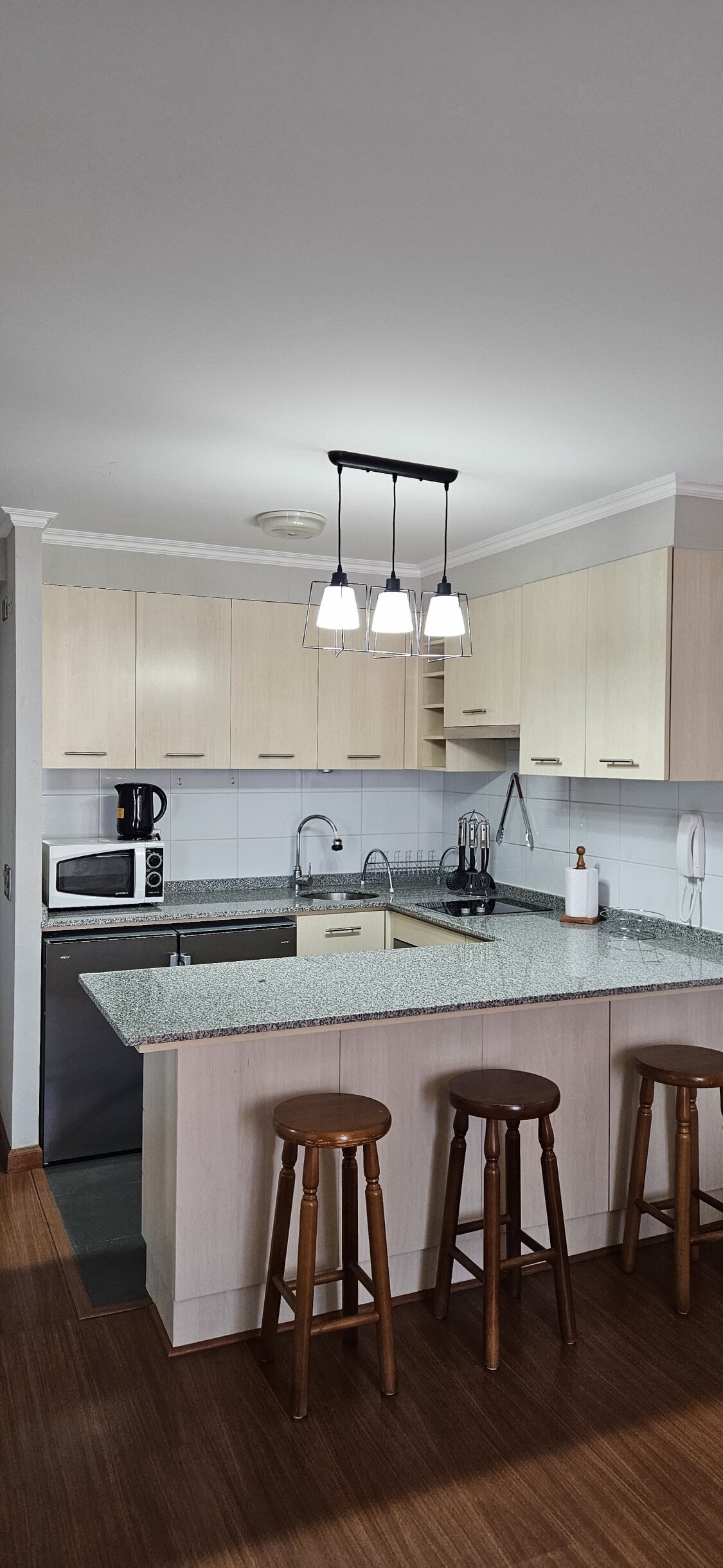 A modern kitchen features light wood cabinetry and a granite countertop, equipped with essential appliances including a microwave and a coffee maker. Three wooden stools are positioned at the countertop, and overhead lighting provides a bright workspace.
