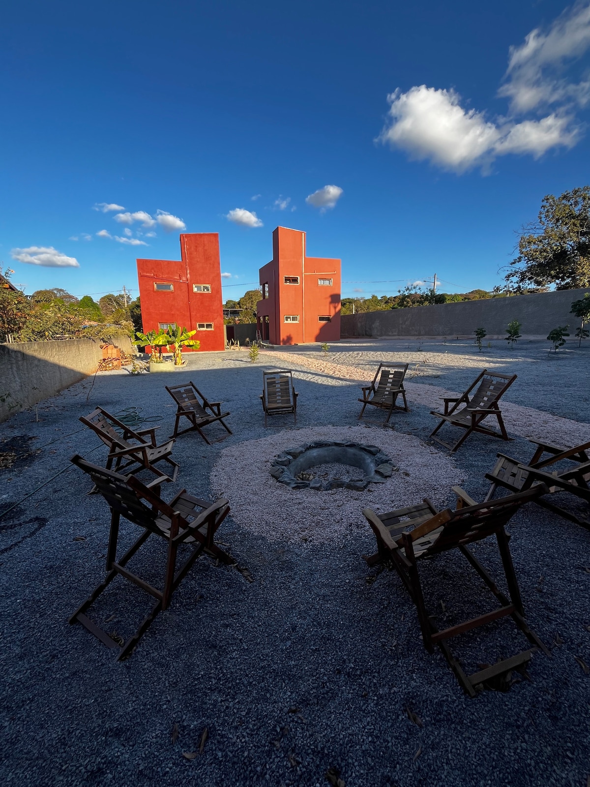 An outdoor gathering area features a circular stone firepit surrounded by several wooden chairs arranged for seating. The backdrop includes two red structures amid a clear blue sky with scattered clouds, set against a landscaped environment.