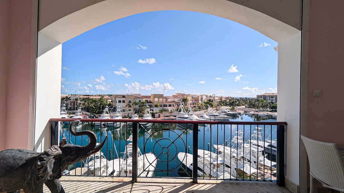 A balcony offers expansive views of the marina, showcasing moored boats and clear blue skies. The railing features an intricate design, while a decorative elephant statue is positioned nearby. Natural light floods the space, enhancing the tranquil scene.