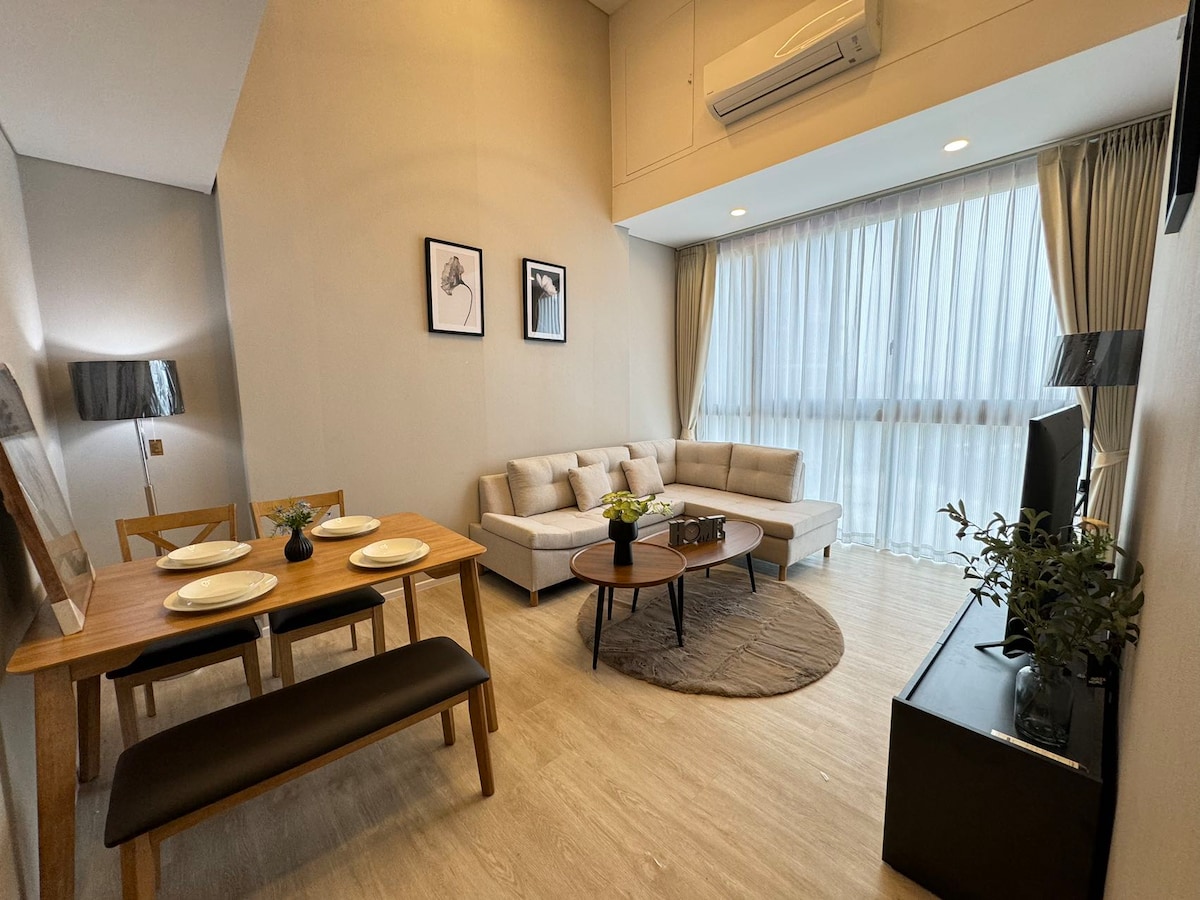 A modern living area features a light-colored sofa arranged around a circular coffee table. A dining table with four plates sits on one side, while large windows are dressed with sheer curtains, allowing soft natural light to fill the space.