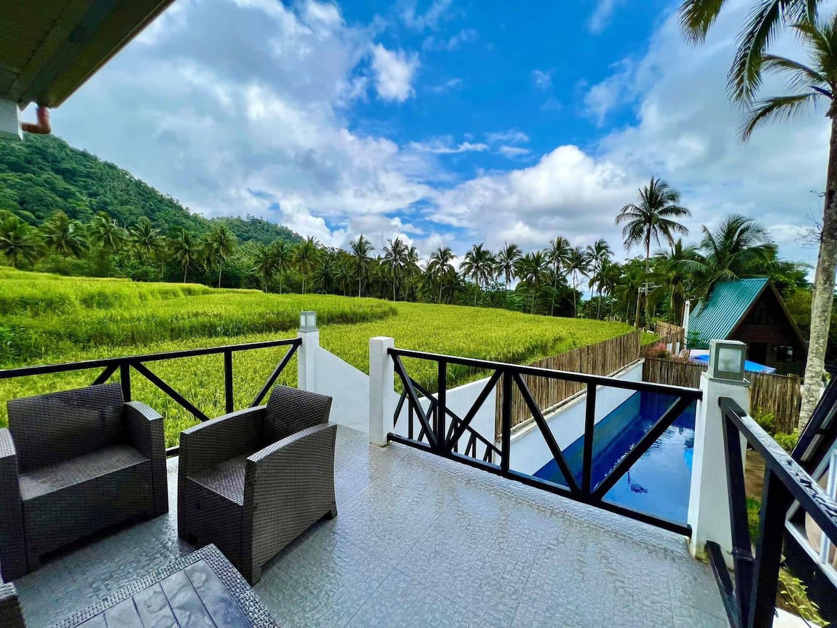 An outdoor balcony is presented with comfortable seating, framed by a view of lush greenery and distant palm trees. The swimming pool is visible nearby, while the sky features a mixture of clouds and blue tones. The tranquil setting offers a sense of peace.