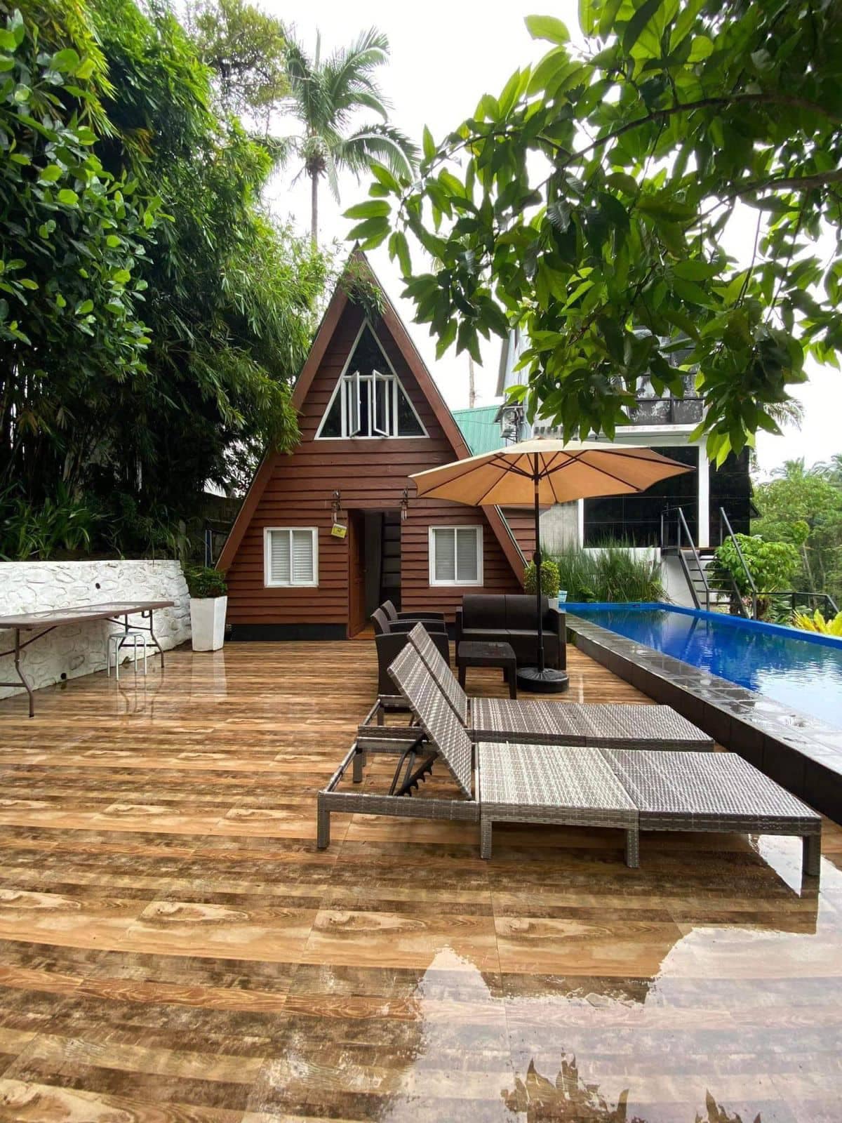 A modern A-frame villa is set against lush greenery, featuring a private infinity pool. Comfortable lounge chairs surround the poolside area, complemented by an umbrella for shade. A wooden patio reflects the serene outdoor environment, enhancing the peaceful atmosphere of the accommodation.