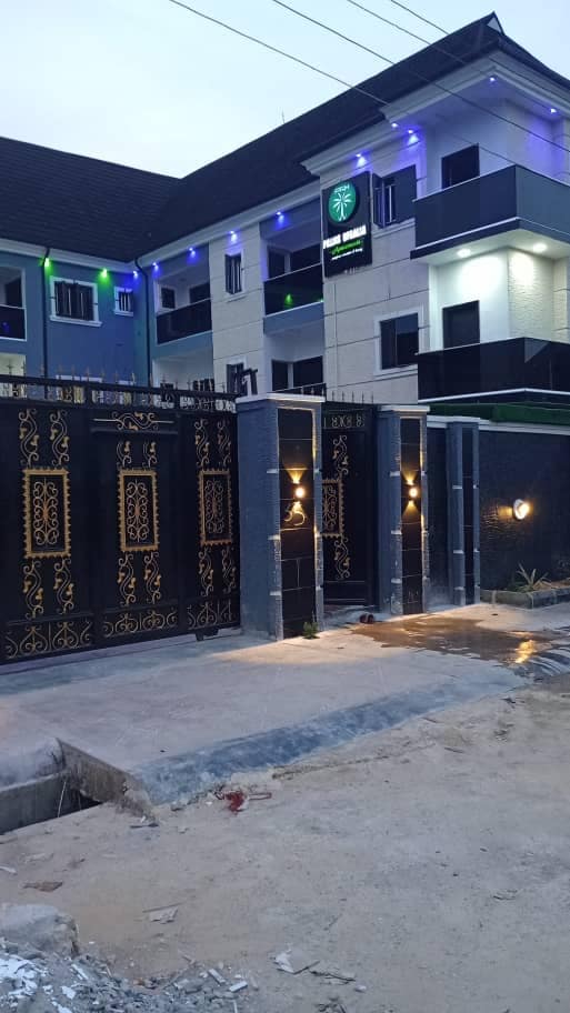 The exterior of the Palms Regalia Luxury Apartment is visible, featuring an ornate metal gate with golden detailing. Soft illumination from wall-mounted lights enhances the entrance. The multi-story building showcases a mix of modern and traditional architectural elements, highlighted by subtle colored lighting.