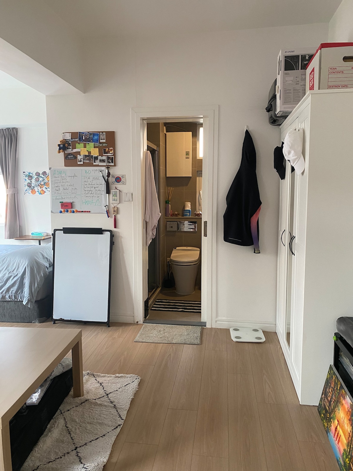 A compact living space is shown, featuring a doorway leading to a bathroom. A whiteboard and bulletin board are mounted on the wall, near a small table and a wardrobe. Soft natural light enters through a window, illuminating the wooden floor.