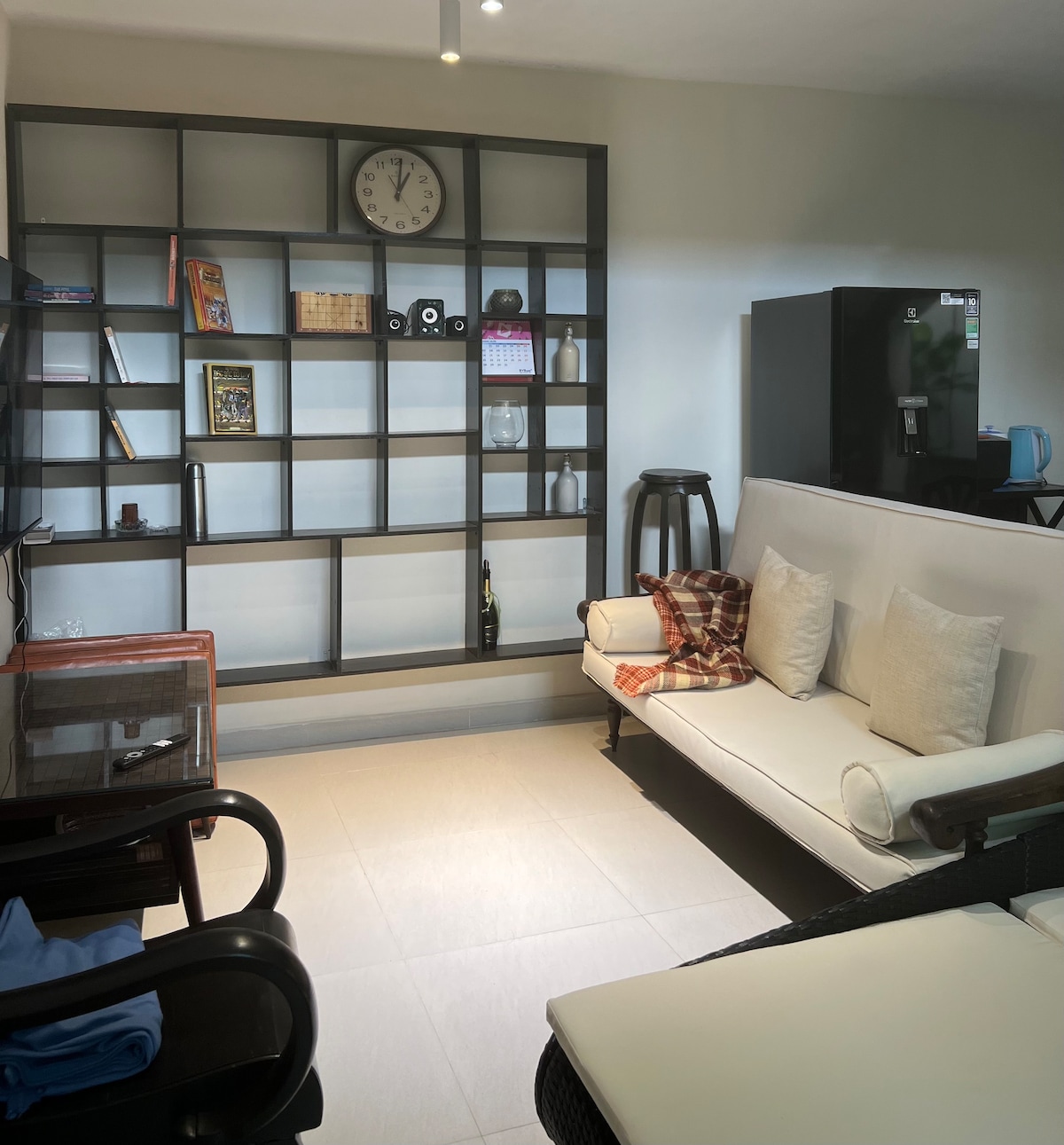 An open living space features a contemporary shelving unit with various decorative items. A clock is mounted on the wall above the furniture, which includes a light-colored sofa adorned with beige cushions and a cozy plaid throw. A dining table is positioned next to a black refrigerator.