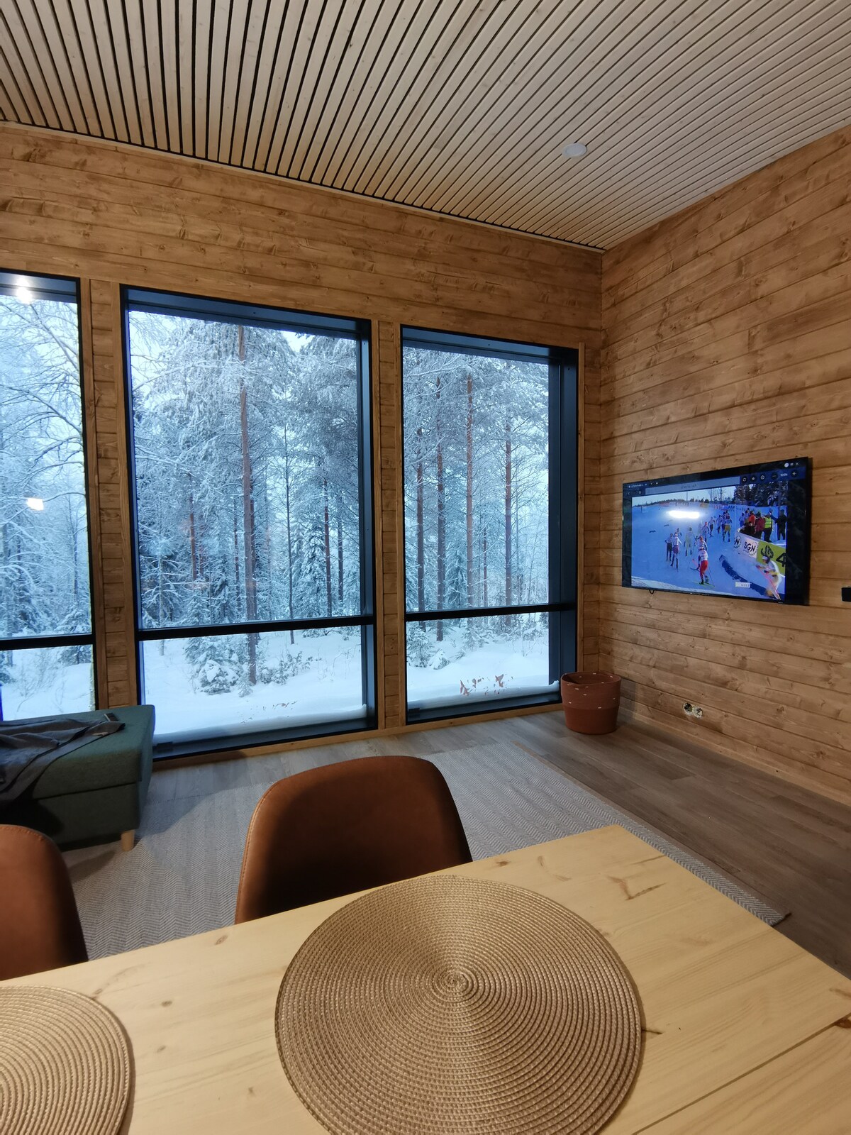 Large windows frame a snowy forest view, allowing natural light to fill the space. A wooden dining table with round placemats is positioned in the foreground, while a comfortable couch offers seating. A flat-screen TV is mounted on the wall, enhancing the inviting atmosphere.