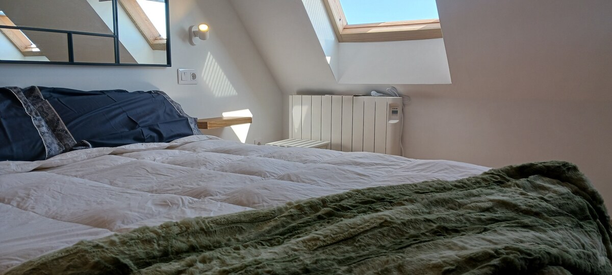 A spacious bed is positioned under skylights, allowing natural light to fill the room. A soft blanket in earthy tones is draped across the bed. A modern radiator is visible against the wall, providing warmth and comfort.