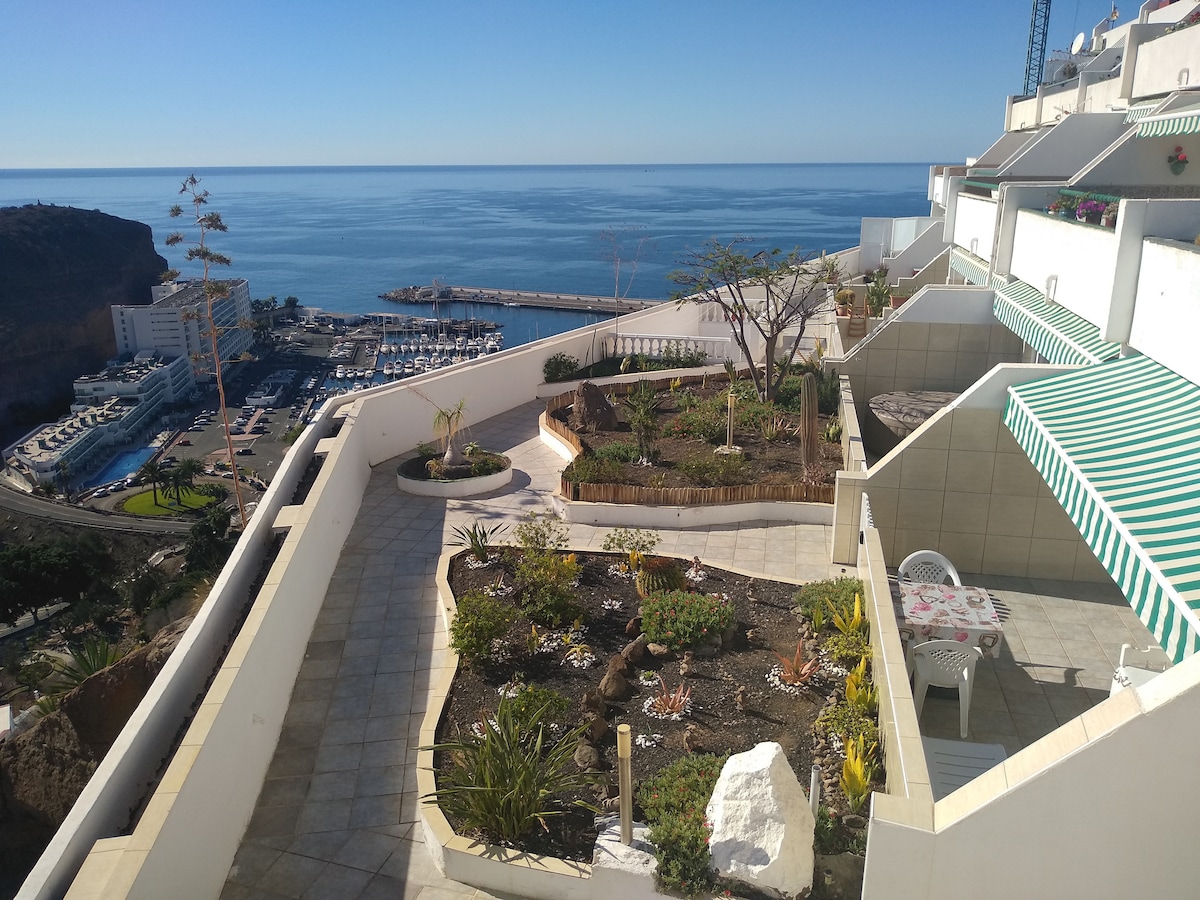 A spacious terrace provides a clear view of the ocean, with a landscaped garden featuring various plants and cactus arrangements. The serene setting includes outdoor seating and vibrant awnings, creating a pleasant environment for relaxation.