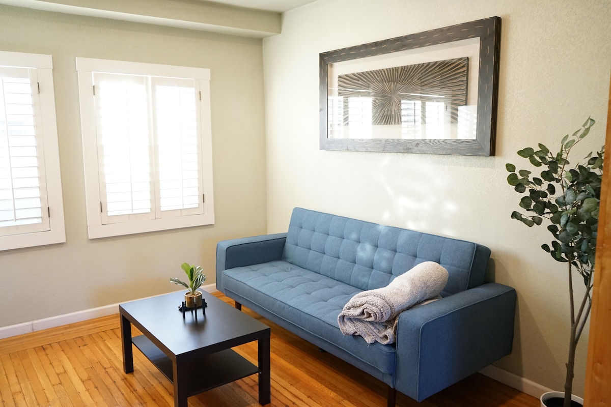 A light-filled living area features a modern, blue sofa accompanied by a black coffee table. A folded throw blanket rests on the sofa, while a small potted plant adds a touch of greenery. The room is complemented by natural light streaming through two shuttered windows.