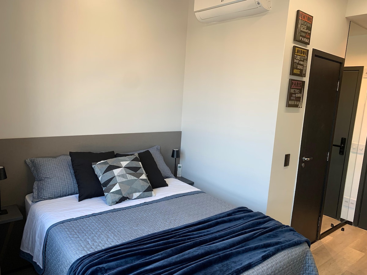 The sleeping area features a spacious bed topped with decorative pillows and a soft throw blanket. Two bedside lamps provide ambient lighting, while textured wall decor adds a modern touch. A door leads to the hallway, and a window offers natural light.