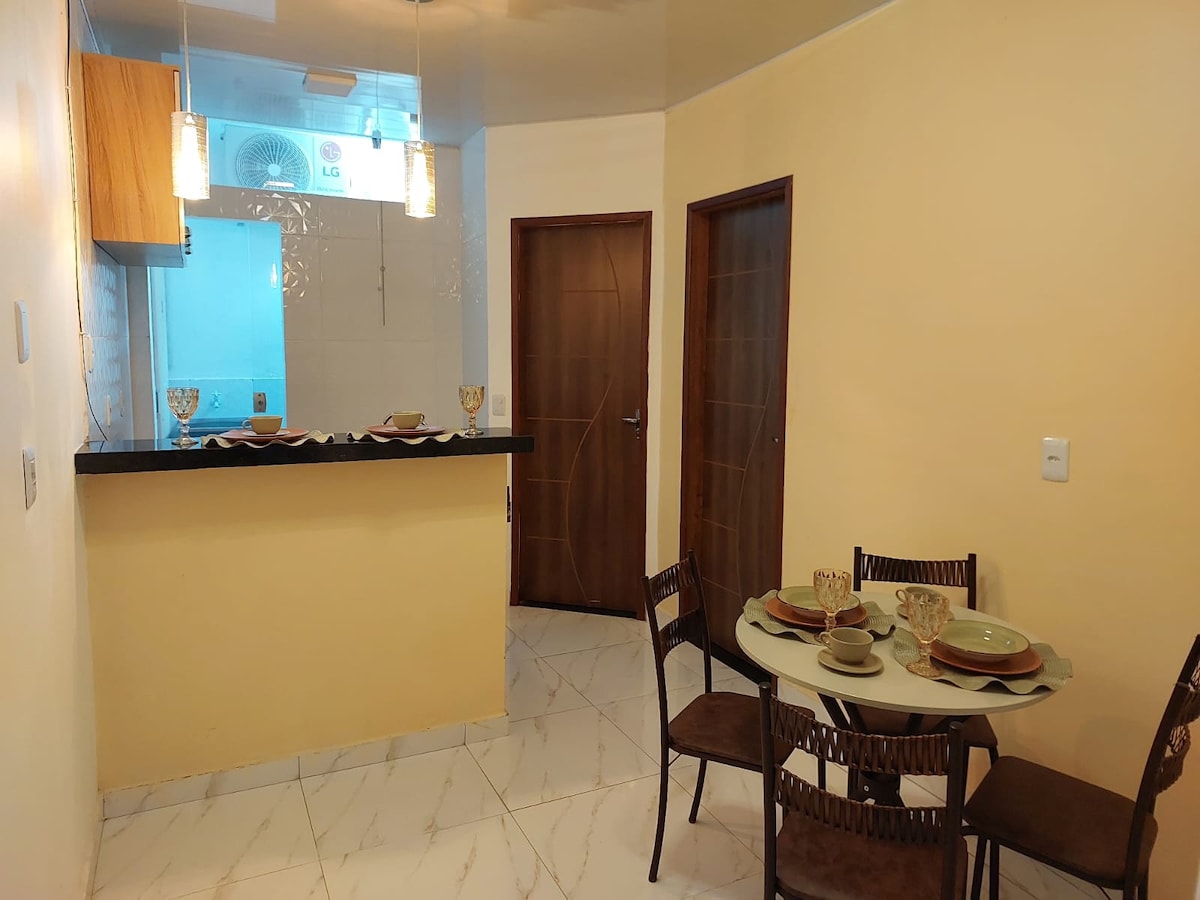 A dining area is visible with a round table set for four, featuring plates and glassware. Soft, warm lighting enhances the space. In the background, a kitchen bar is partially seen, complemented by light-colored cabinetry and a tiled backsplash.