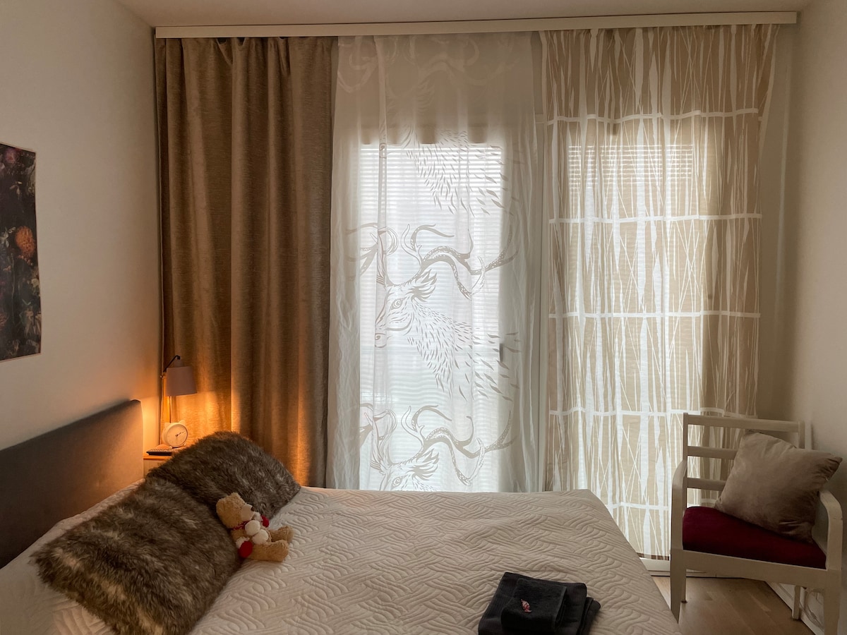 A cozy sleeping area features a spacious bed with a textured white coverlet. A soft throw and a teddy bear are positioned on the bed. Nearby, a chair with a red cushion is visible. Natural light filters through sheer curtains, creating a peaceful ambiance.