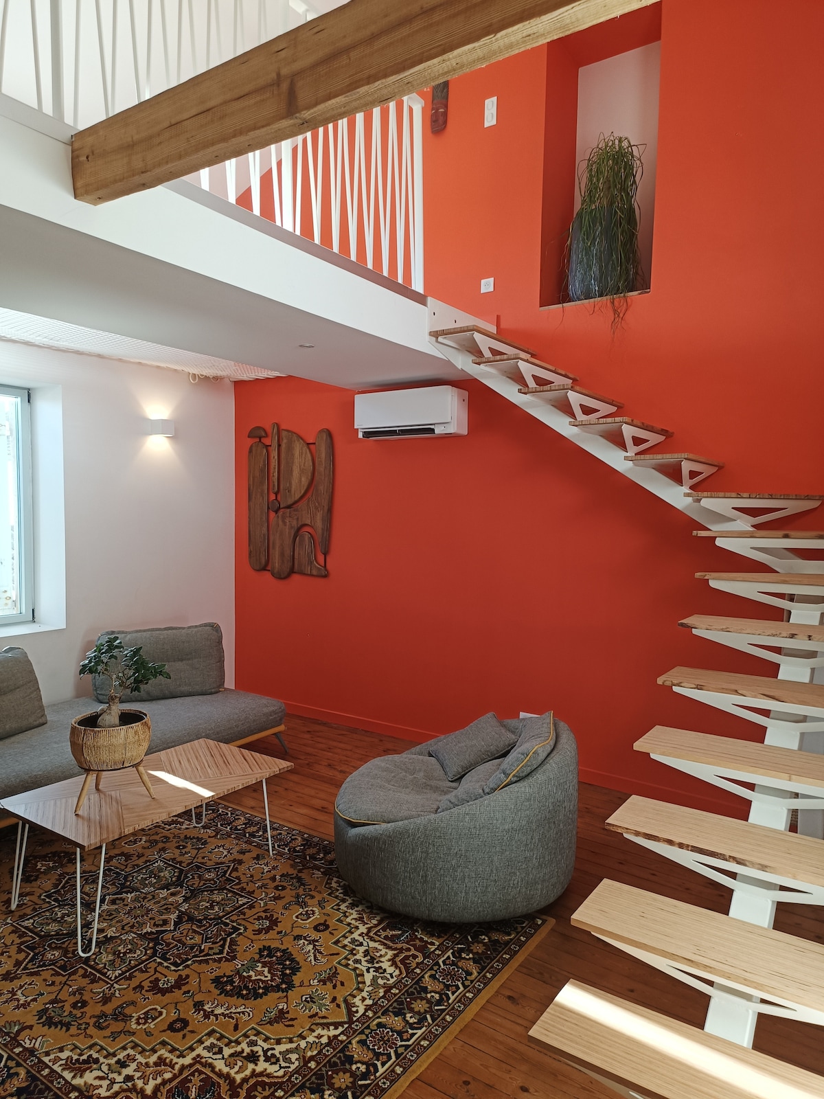 A two-level living space features a striking orange accent wall, complemented by a wooden beam and a spiral staircase. Comfortable seating is arranged on a patterned rug, while a potted plant adds a touch of greenery. Natural light streams through large windows, illuminating the area.