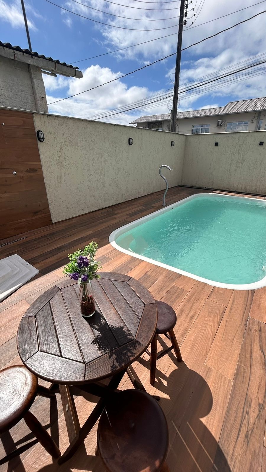 An outdoor area features a small round table with two wooden chairs positioned beside a private swimming pool. The pool, surrounded by wooden decking, reflects sunlight under a clear blue sky. A vase with flowers adds a touch of color to the setting.