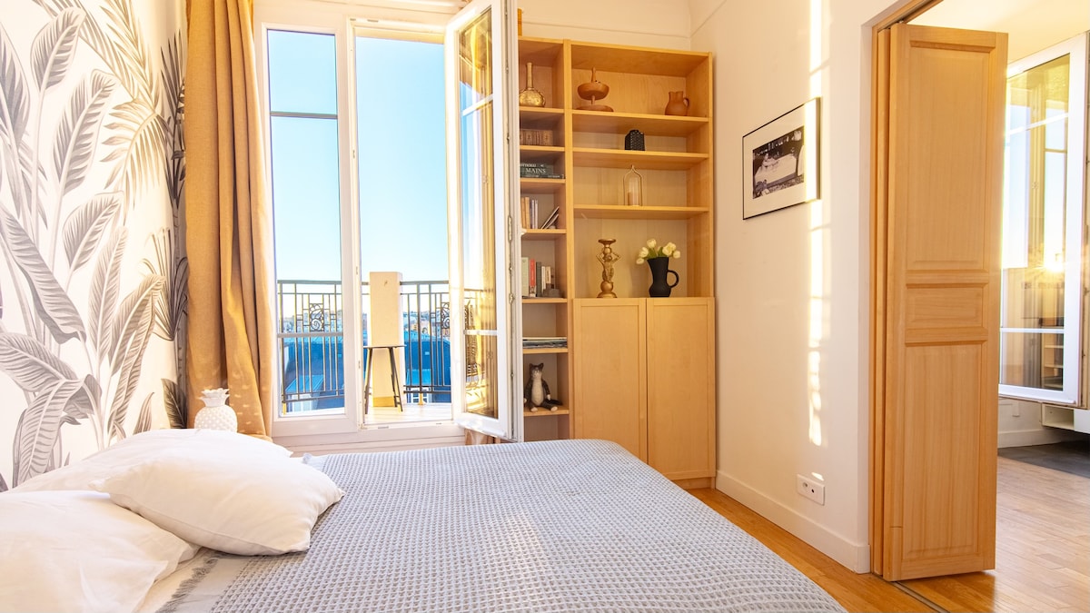 A bright sleeping area features a spacious bed with white pillows. Large windows open to a balcony that offers a view. A wooden bookshelf is positioned against the wall, complemented by decorative items. Sunlight fills the room, enhancing the light-colored decor.