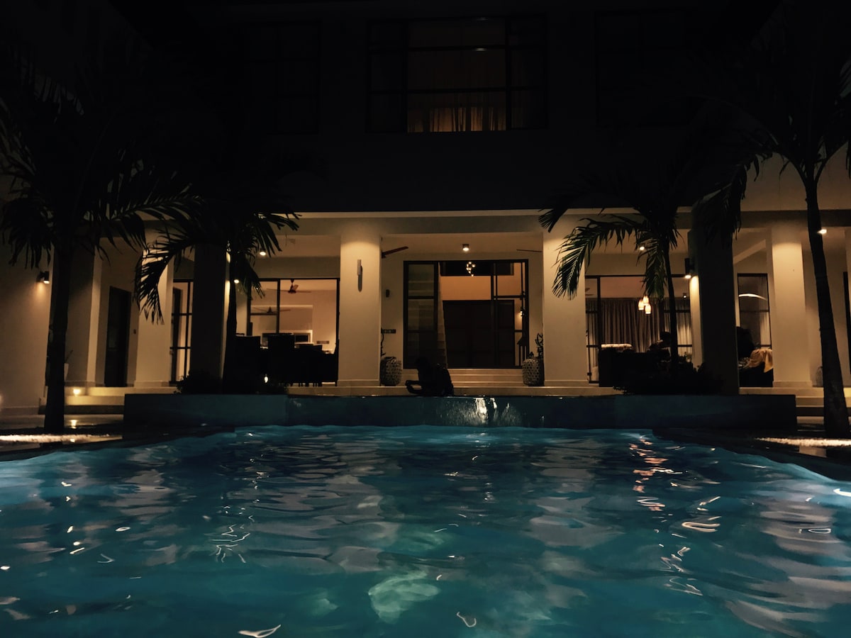 A tranquil pool is illuminated under the warm glow of surrounding lights, reflecting the serene atmosphere of the outdoor space. The villa's spacious entrance is visible in the background, framed by elegant palm trees enhancing the inviting ambiance.