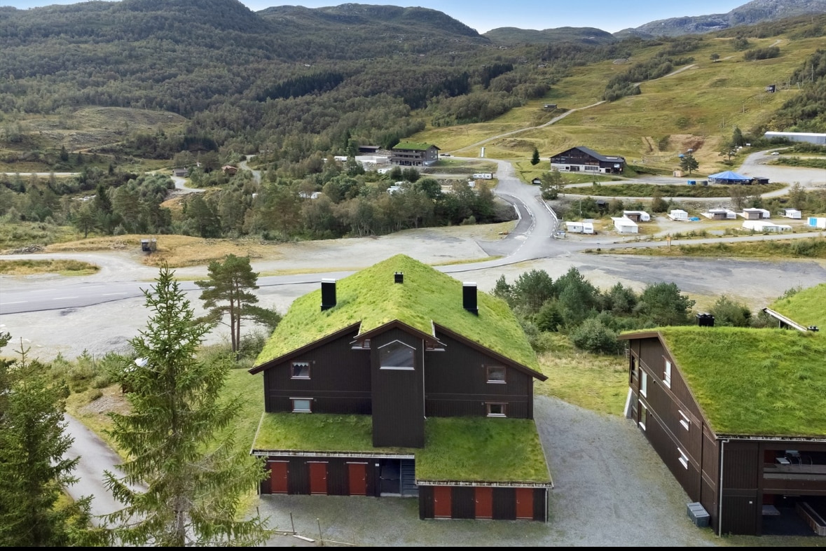 An aerial view depicts a brown mountain lodge with a green, grass-covered roof, surrounded by a natural landscape of hills and trees. Nearby structures and winding roads are visible, indicating a tranquil and rustic setting.