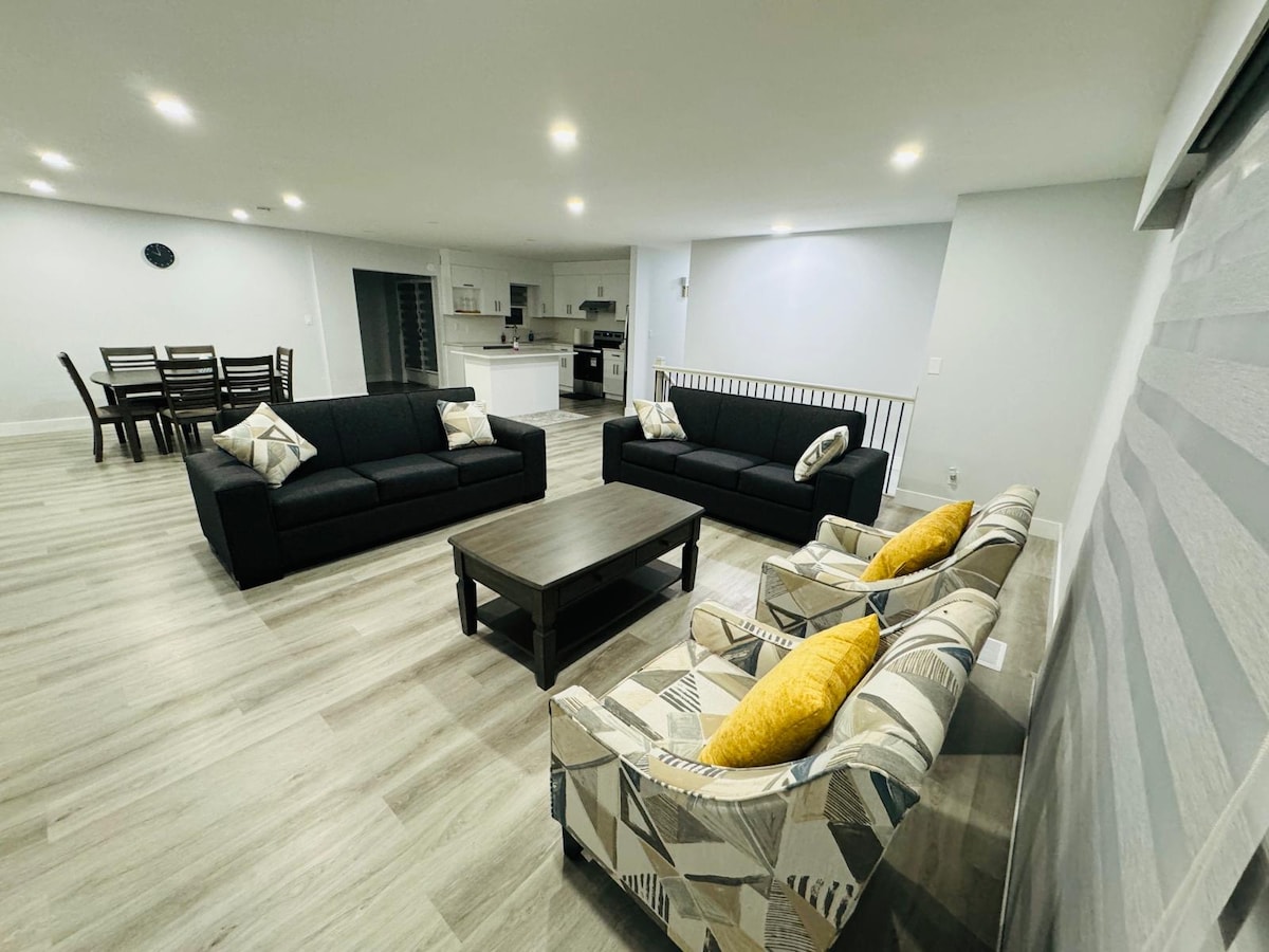 A spacious living area features two sleek black sofas and a patterned armchair with yellow cushions. A wooden coffee table is centered between the seating, while a dining area with a dark wood table and chairs is visible in the background. Soft lighting enhances the clean, modern aesthetic.