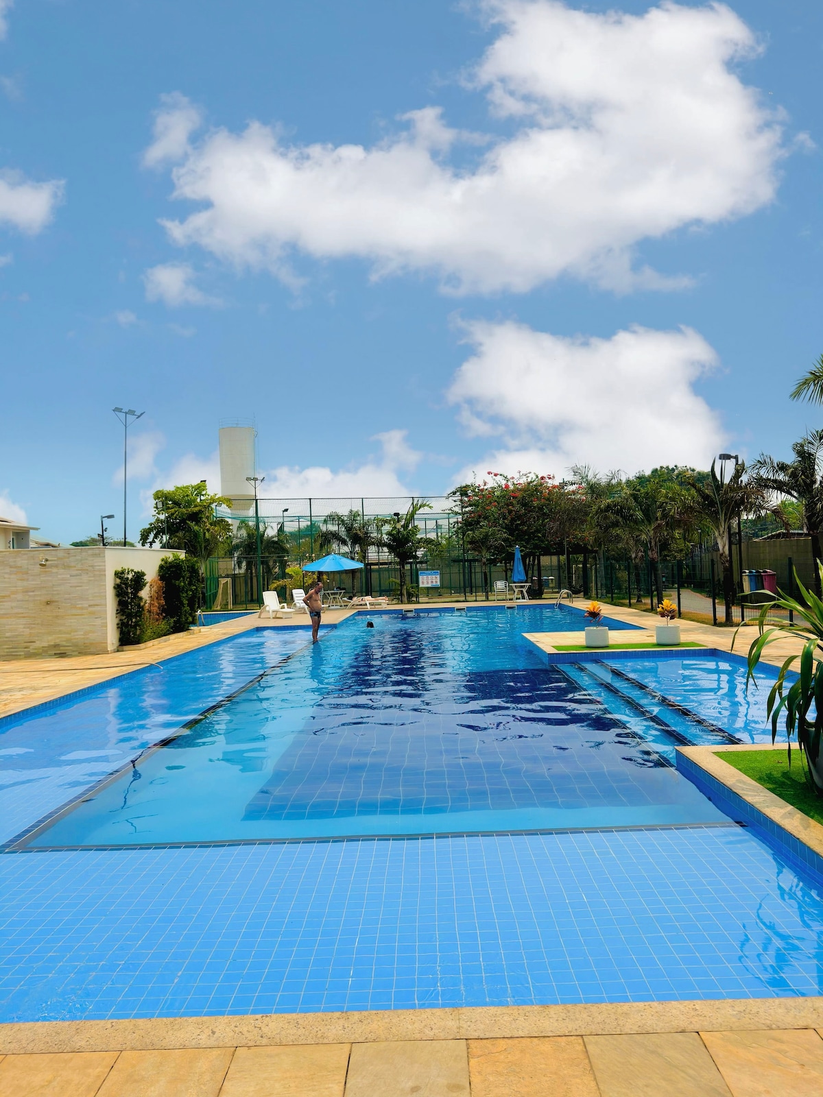 A long swimming pool with clear blue water is surrounded by lush greenery and palm trees. The pool area includes lounge chairs and is set against a backdrop of light-colored buildings and a clear sky with fluffy clouds.