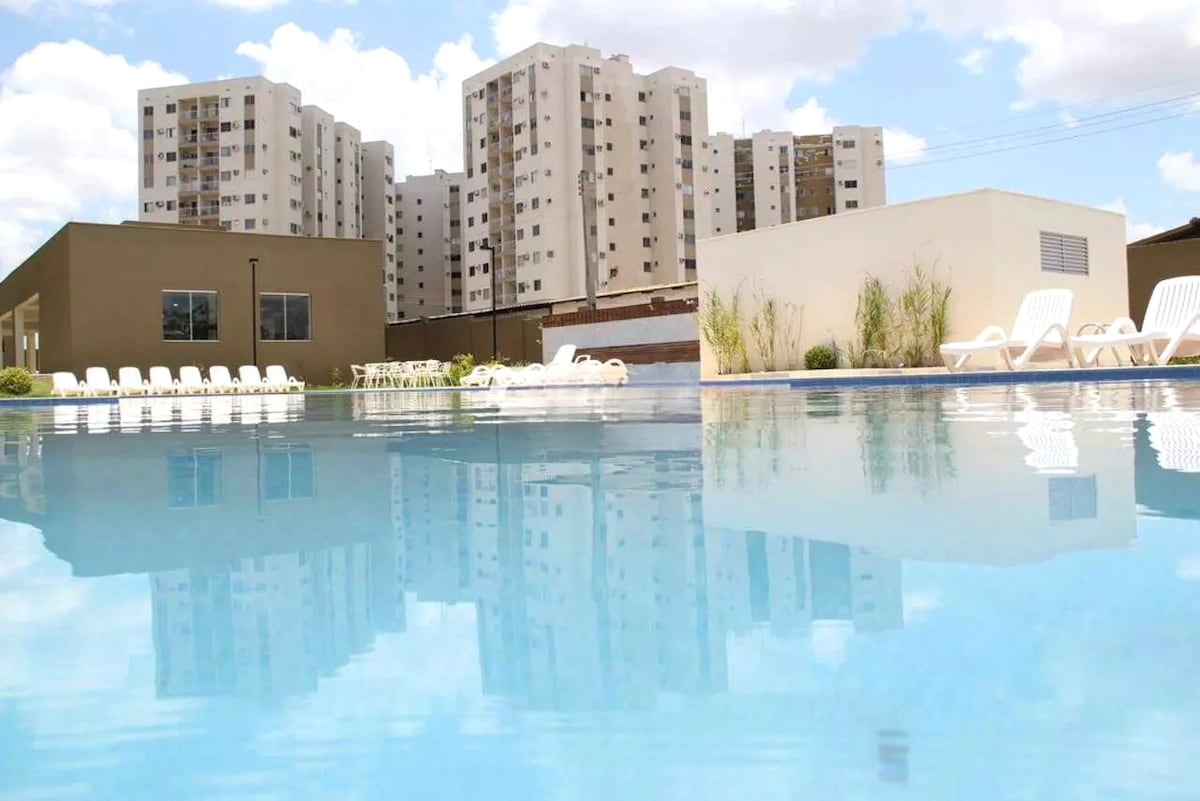 A spacious swimming pool is seen reflecting the surrounding buildings and sky. Lounge chairs line the pool's edge, providing ample seating space. The serene water creates a calming focal point amidst the vibrant, urban backdrop.