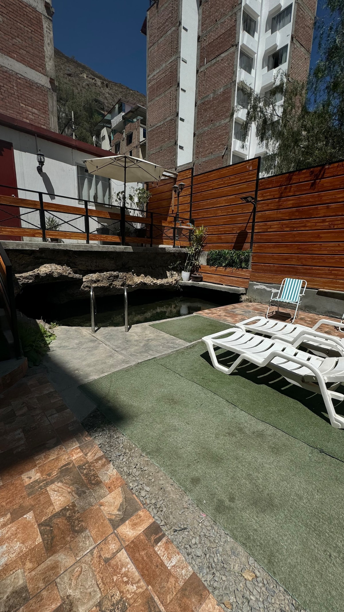 An outdoor area features a small thermal pool surrounded by stone elements. Sun loungers are arranged on a green lawn, with an umbrella providing shade. Wooden fencing lines the perimeter, enhancing the quiet ambiance of the space.