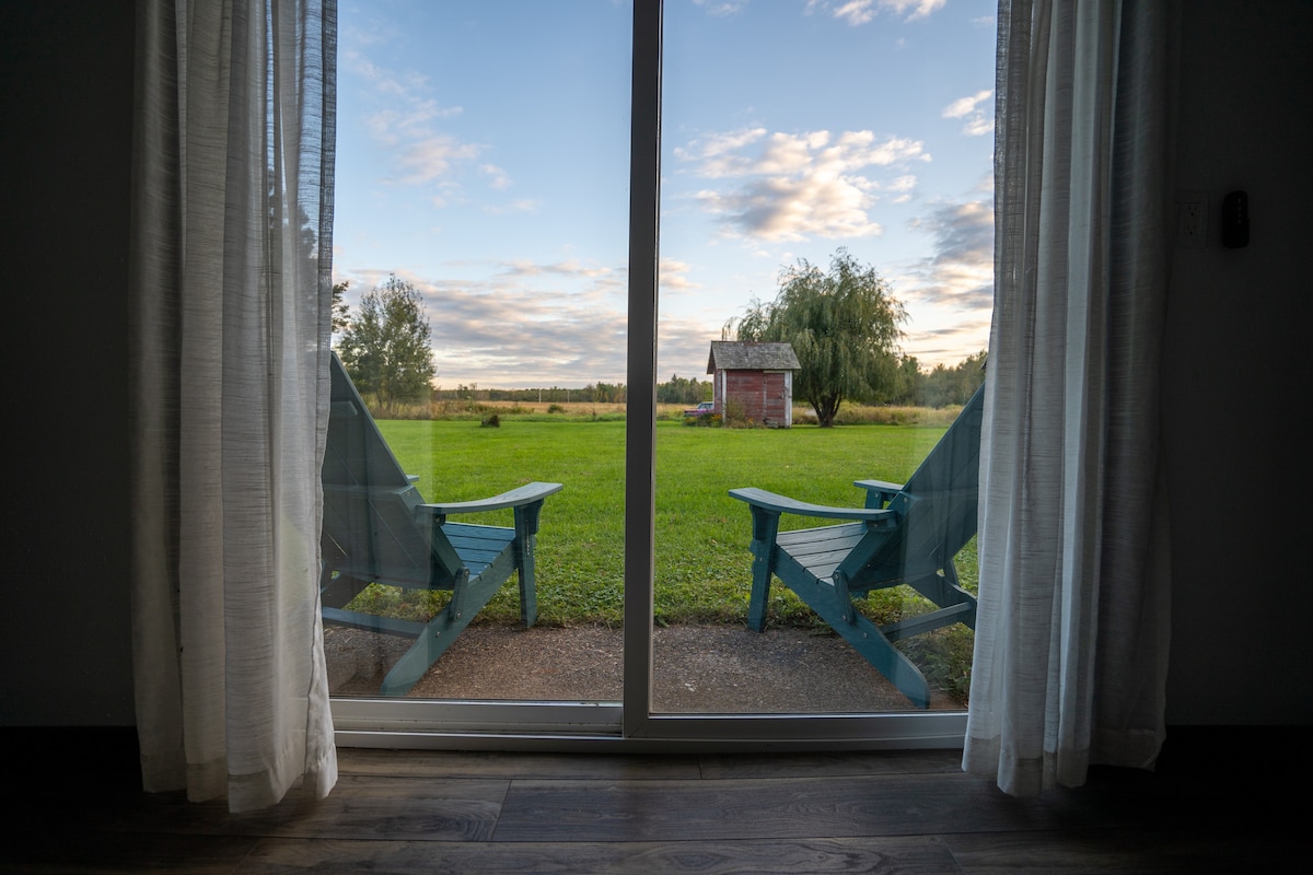 Two blue Adirondack chairs are placed on a patio, facing an expansive green yard. The view includes a small barn in the distance under a sky adorned with scattered clouds, and light curtains frame the glass sliding doors.
