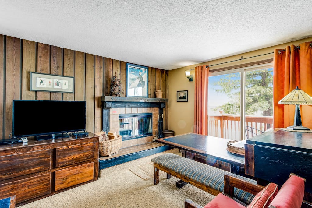 A cozy living area features a wood-burning stove set beneath a rustic mantel. A television is positioned on a wooden console. Large windows provide natural light and access to a balcony. Wooden accents and comfortable seating enhance the relaxed atmosphere.