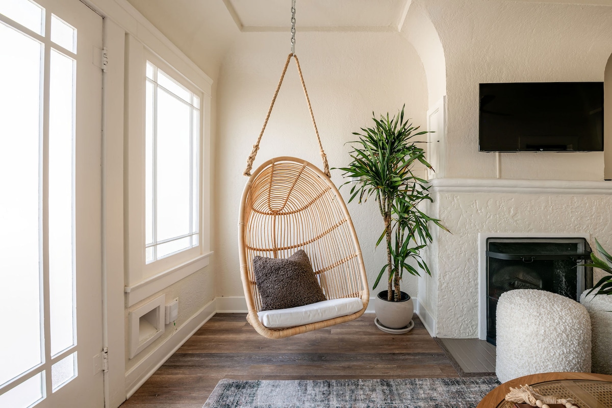 A hanging basket chair is suspended in a cozy corner, complemented by a plush cushion. A green potted plant adds a vibrant touch, while a flat-screen television is mounted on the wall. Natural light filters in through nearby windows, enhancing the inviting atmosphere.