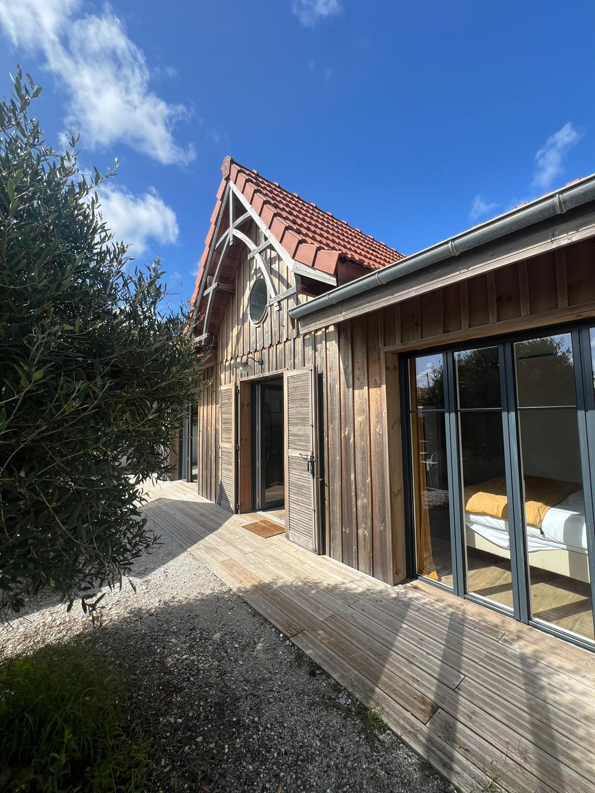 The exterior of the house is showcased, highlighting a wooden facade and a sloped roof. Large windows reflect natural light, and a wooden pathway leads to the entrance. Surrounding greenery adds a touch of nature, creating a harmonious blend with the architecture.