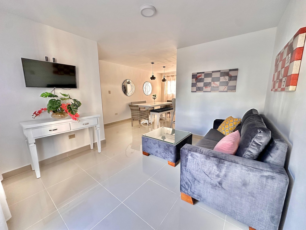 A clean and modern living area is featured, with a gray sofa accompanied by decorative cushions. A glass coffee table sits in front, and a flat-screen television is mounted on the wall. A dining space is visible in the background, showcasing a round mirror and a contemporary dining table.