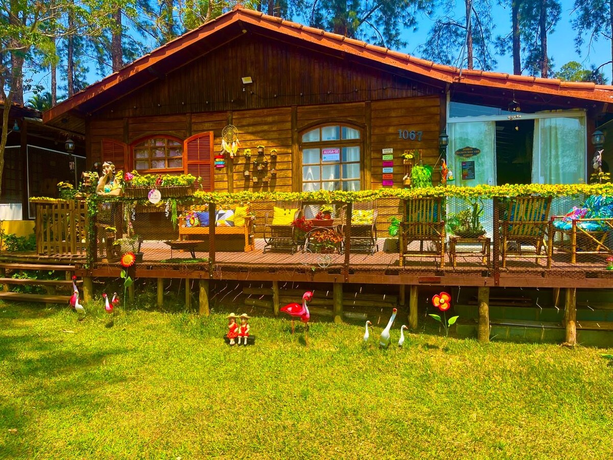 A charming wooden cabin is presented, featuring a welcoming porch adorned with colorful cushions and decor. The surrounding lawn is enhanced by playful garden ornaments, including flamingos and flowers, set beneath tall trees that provide a sense of serenity.