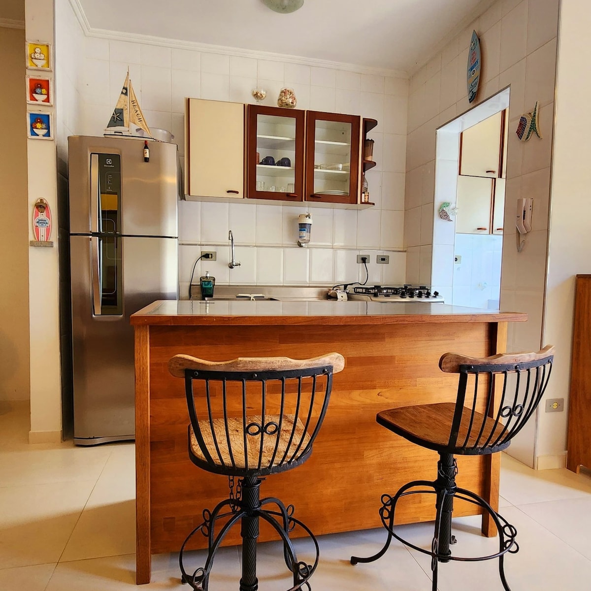 A functional kitchen space is showcased, featuring modern appliances, including a stainless steel refrigerator and a gas stove. Wooden bar stools are positioned at the kitchen counter, which integrates seamlessly with the bright and airy atmosphere. Cabinets are adorned with decorative elements reflecting a coastal theme.