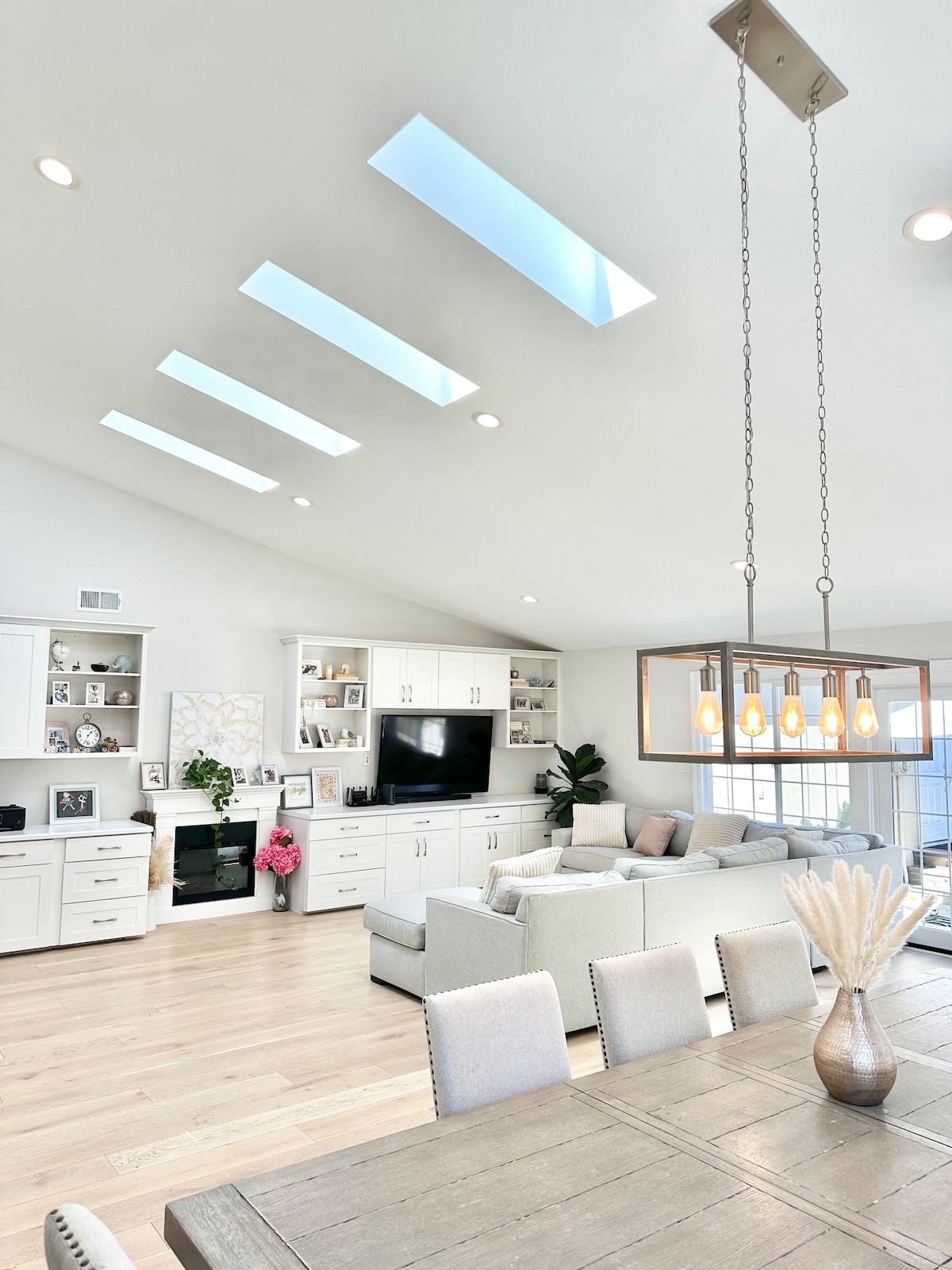 An airy living room features a high ceiling with skylights, allowing natural light to fill the space. A light-colored sectional sofa is positioned near a wall unit with a television. A dining area with a wooden table and upholstered chairs is visible in the foreground.