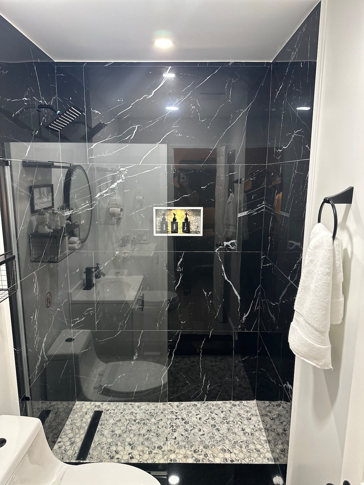 A modern bathroom features sleek black marble walls accented with white veins. A glass walk-in shower is visible, equipped with a rainfall showerhead. A round mirror reflects the space, while plush towels hang neatly. A pebble-stone floor complements the overall contemporary design.