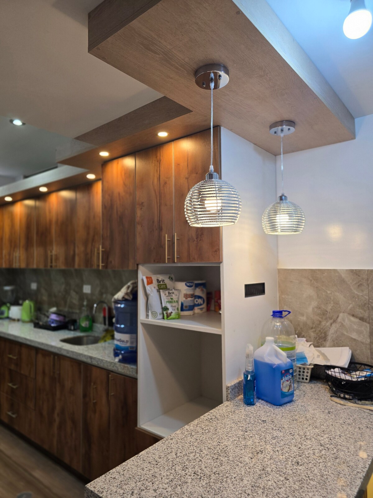 The kitchen features wooden cabinetry, complemented by pendant lights. A granite countertop provides ample space for preparation, while appliances and kitchenware are neatly arranged. A small nook is visible for storage, ensuring efficient organization within the space.