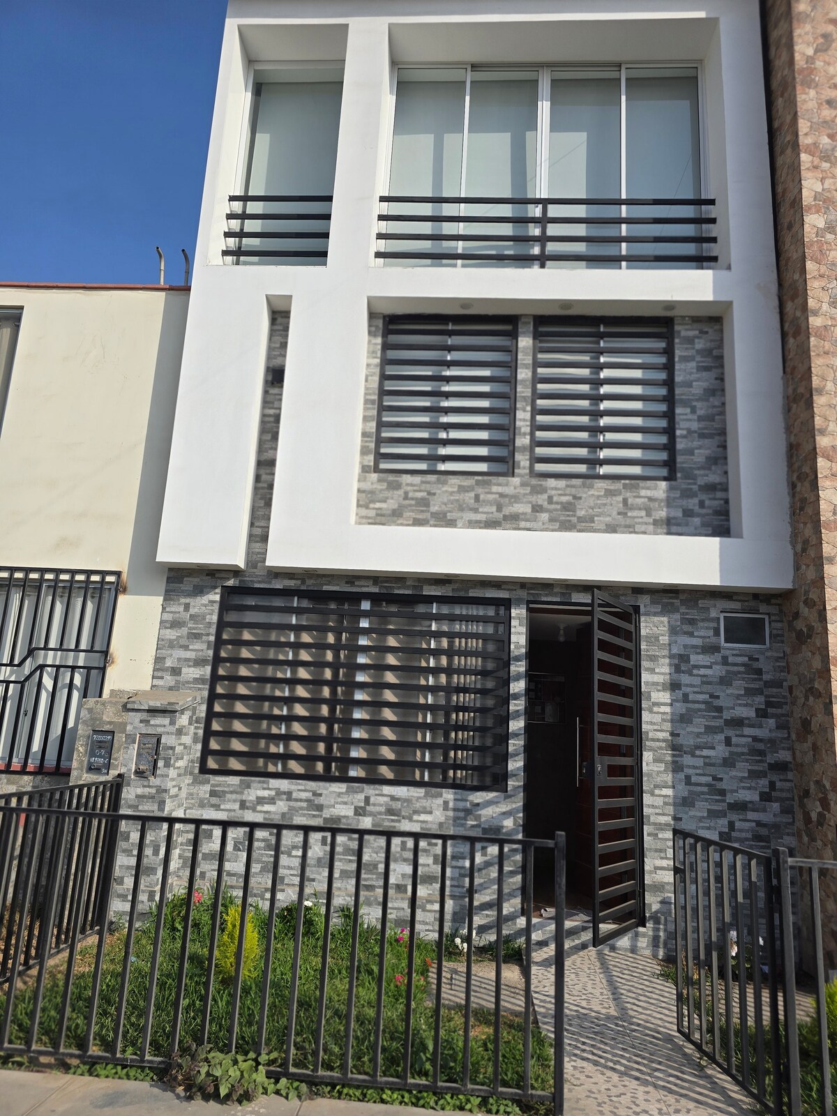 The exterior of a modern, three-story building is presented, featuring a combination of stone and glass elements. Large windows allow natural light to enter, while sleek black railings provide a contemporary touch. A well-maintained garden with greenery is visible in front of the entrance.