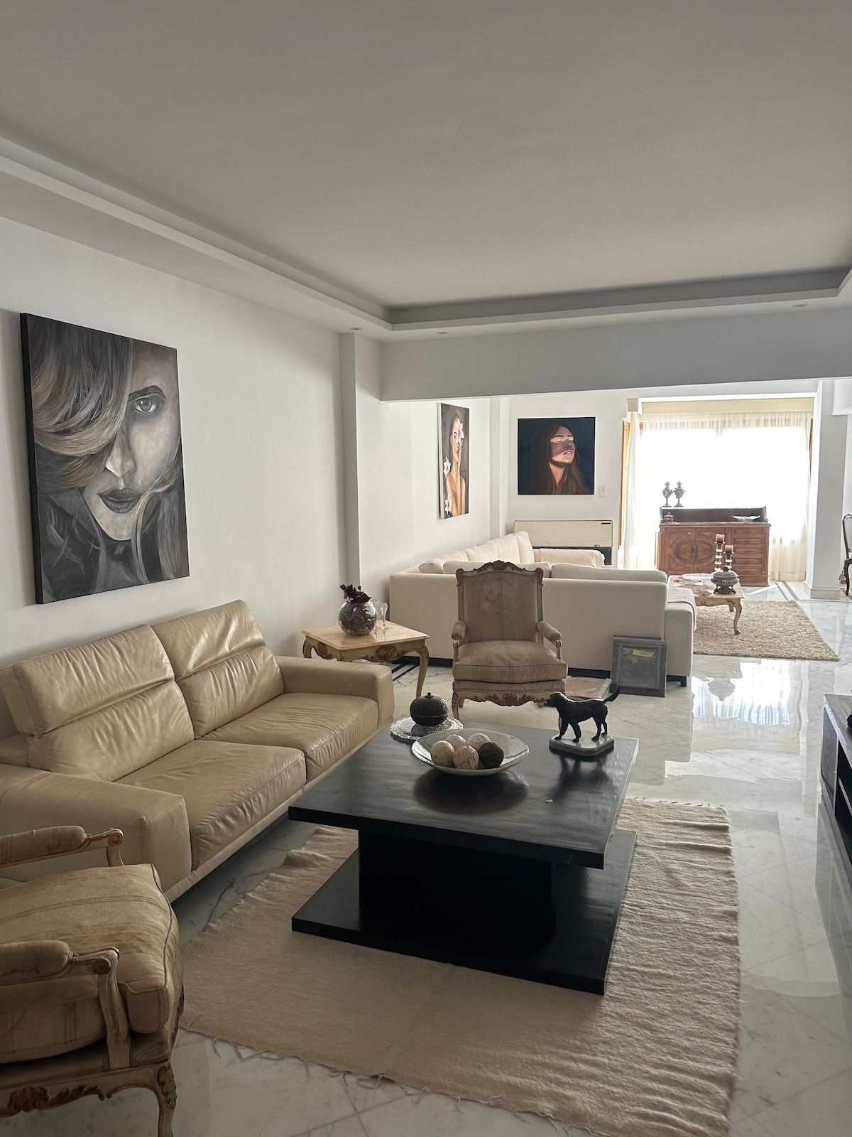 A sunlit living room is decorated with neutral tones and elegant furnishings. A plush cream sofa and a wooden coffee table are positioned on a soft area rug. Decorative artwork adorns the walls, while a large window offers natural light and a view of the outdoors.