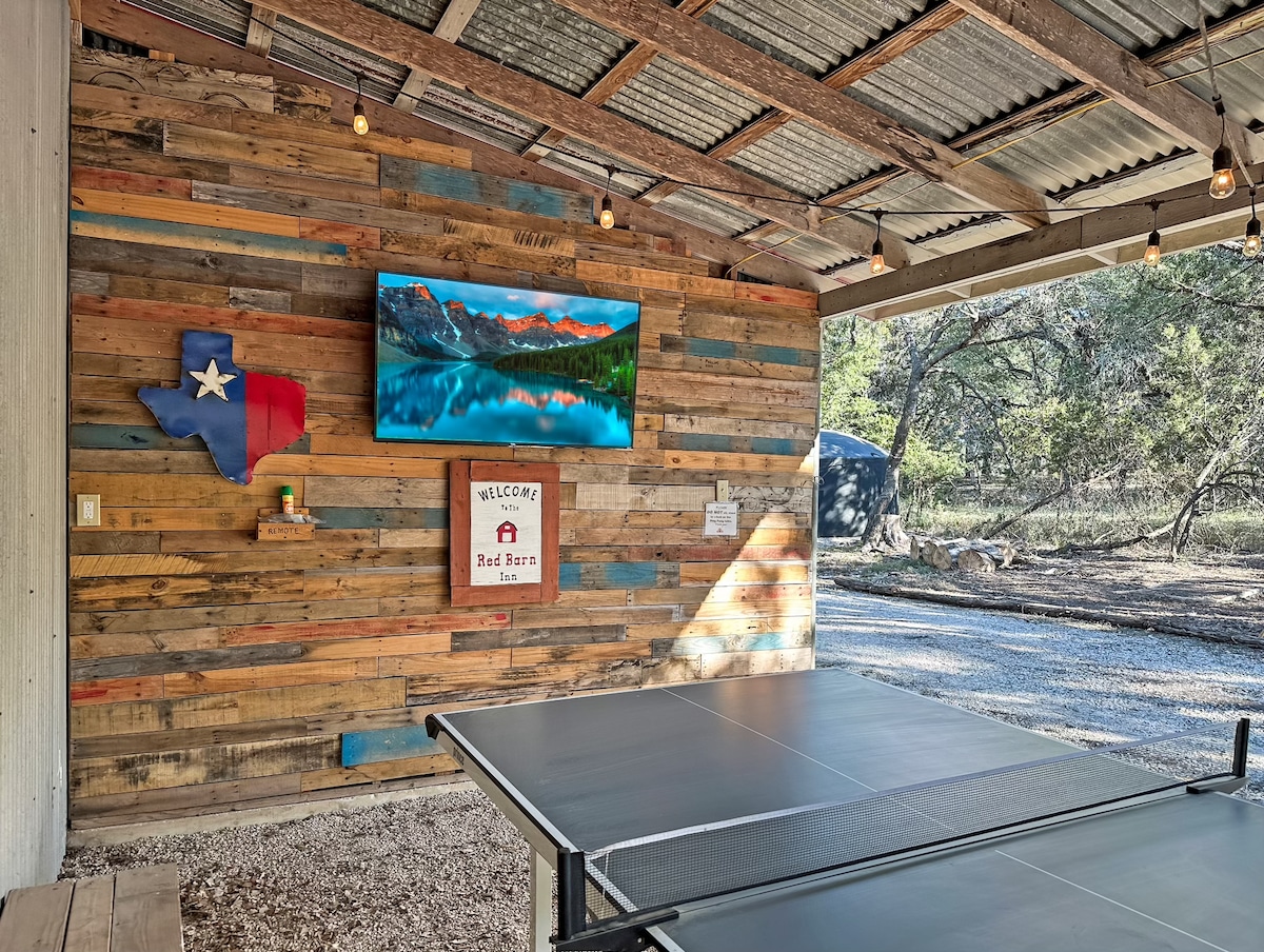 An outdoor entertainment area features a ping pong table beneath a rustic wooden roof. A Texas-themed wall decor is displayed alongside a mounted television. Warm lighting is provided by string lights, creating a cozy environment surrounded by nature.