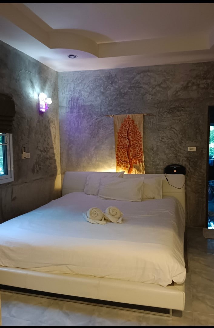 A spacious bed with crisp white linens is featured in a calming room, complemented by textured gray walls. A decorative fabric art piece with a tree design hangs above the bed, while soft lighting creates a soothing atmosphere. A window provides a view of the outdoors.
