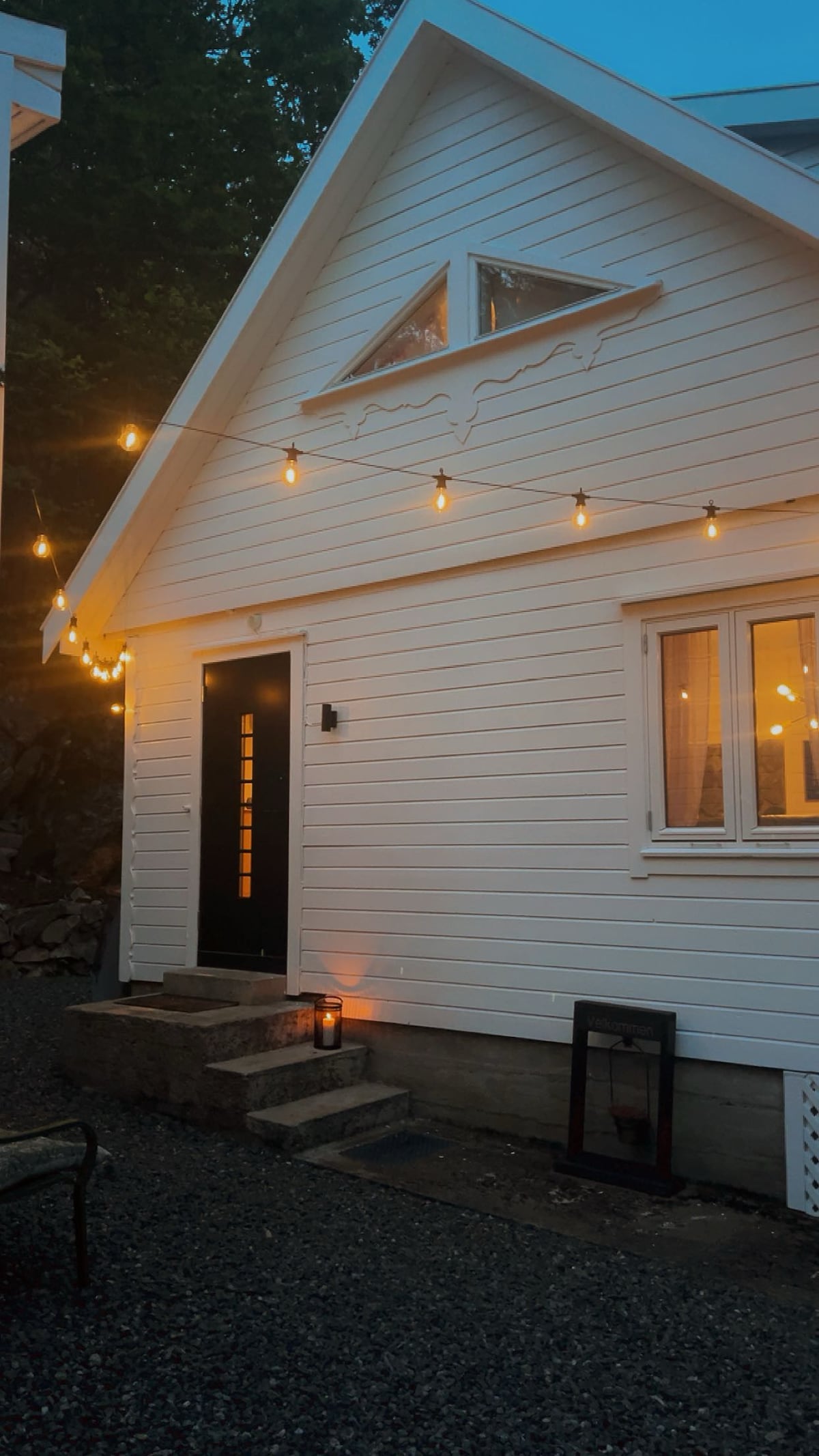 The exterior of the charming cabin is seen, featuring a white, gabled façade with lighted string decorations. A black front door is framed by a window, providing a warm glow. Gravel pathways lead to the entry, complemented by soft lighting from a lantern and outdoor fixtures.