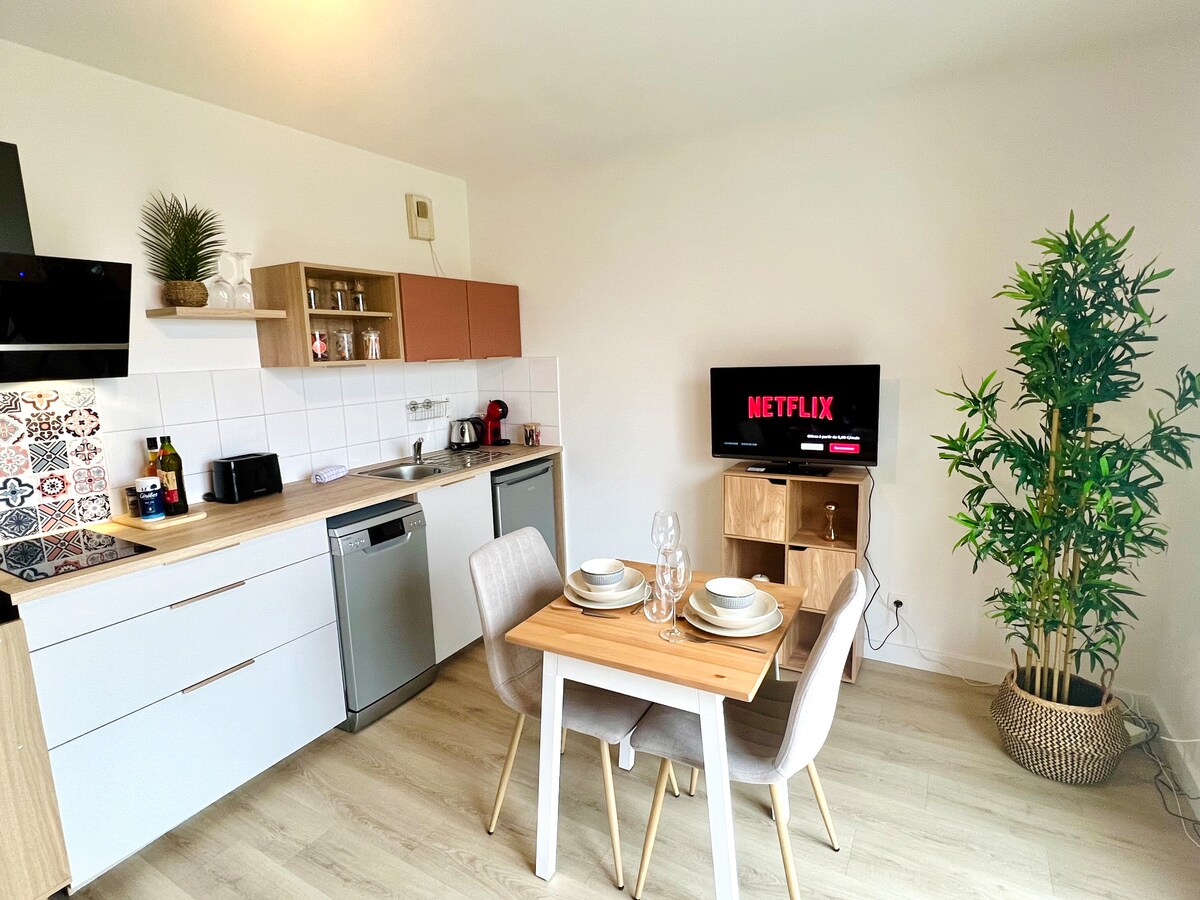A modern kitchen area features light wood cabinetry and a spacious dining table set for two. Kitchen appliances, including a dishwasher and coffee maker, are clearly visible. A small entertainment unit displays a television with Netflix, alongside a decorative plant providing a touch of greenery.