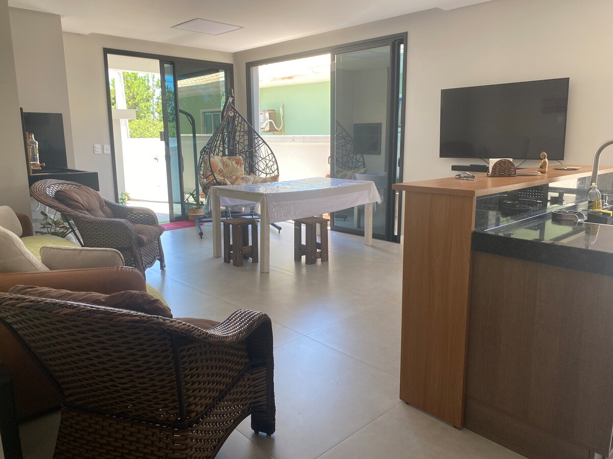 An open living and dining area features large glass doors that invite natural light. A dining table is set between a cozy seating arrangement, which includes a hanging chair. A kitchen area is visible with modern appliances and a contrasting countertop.