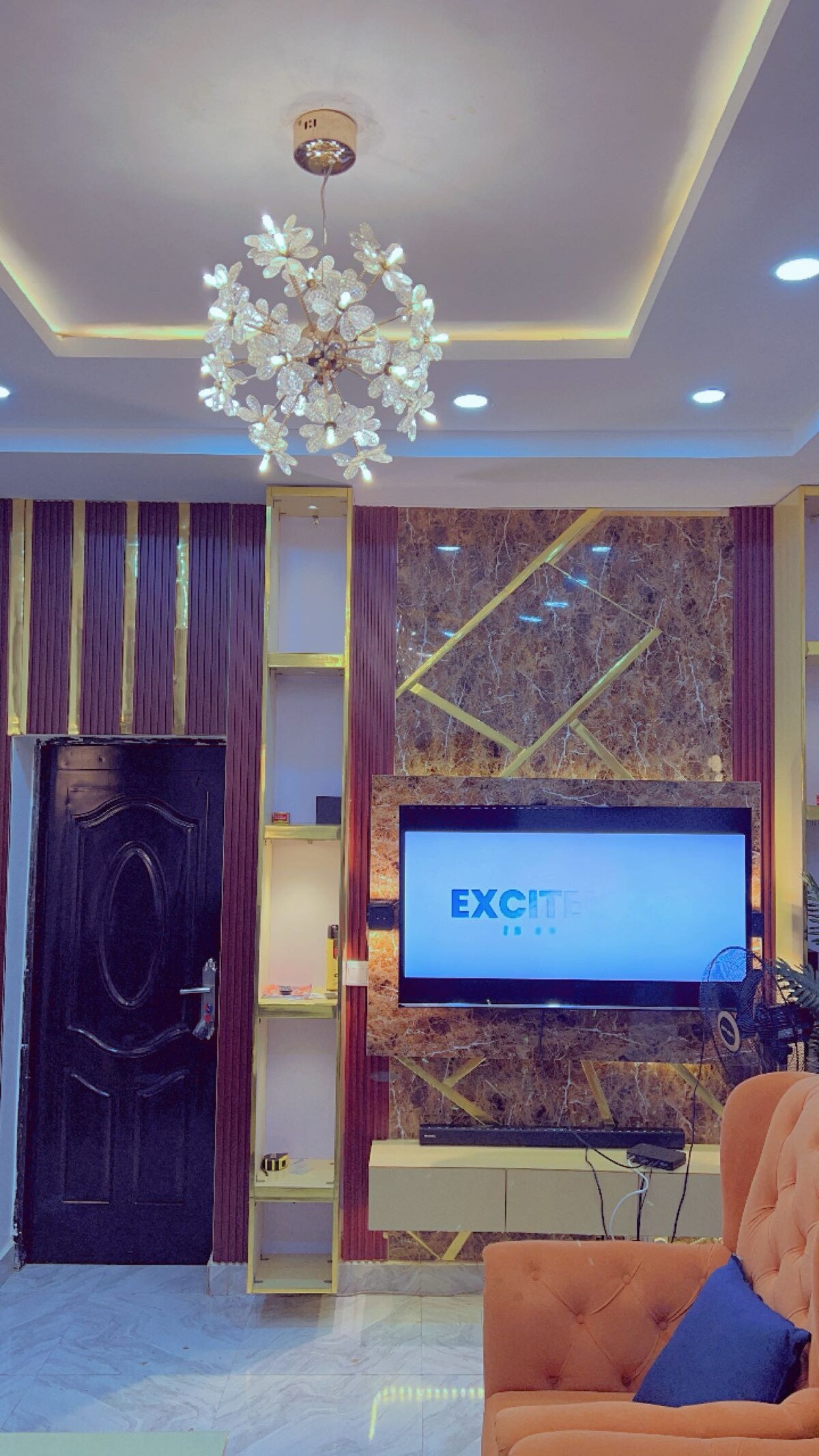 A modern living space is showcased with a stylish chandelier featuring floral accents. A sleek television is mounted on a decorative wall, complemented by ambient lighting. The room is furnished with a comfortable seating area, designed for relaxation and enjoyment.