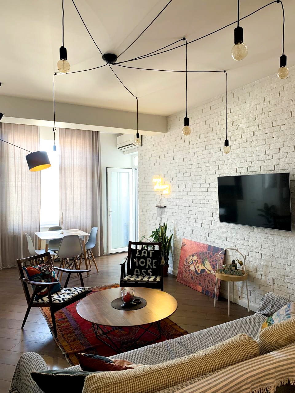 An inviting living area features a stylish sofa and a round coffee table, surrounded by modern pendant lighting. A large wall-mounted television is positioned against a textured white brick backdrop, while a dining table with chairs is visible in the background. Decorative elements include framed artwork and a plant.